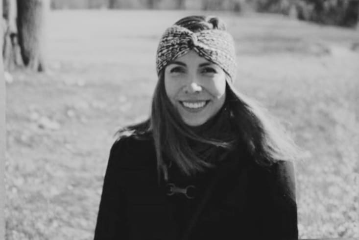 Irene Preciado, Directora Creativa de Grupo en México y parte del programa Líderas de VML, aparece en esta imagen en blanco y negro. Es una mujer con cabello oscuro largo y una sonrisa amable, luce una diadema anudada con un patrón y un abrigo oscuro con un broche. El fondo, ligeramente desenfocado, muestra un entorno natural con follaje y un tronco de árbol.