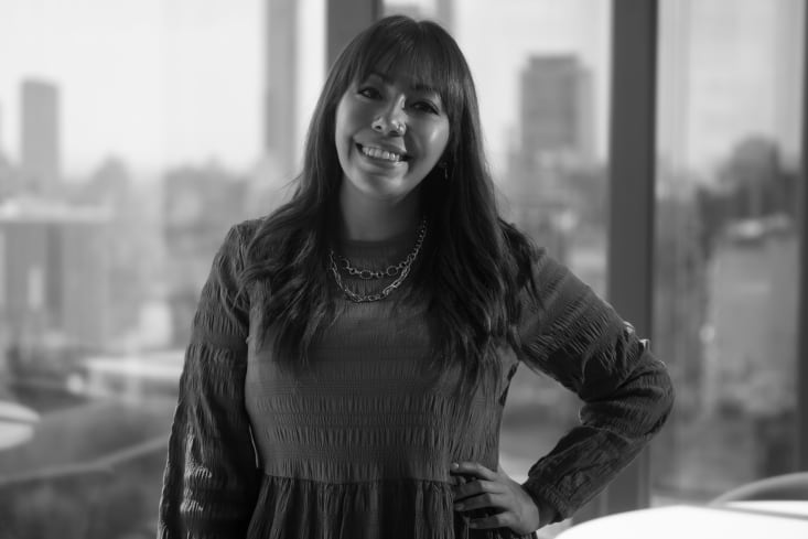 Montserrat Diaz, miembro del programa Líderas de VML, aparece en esta imagen en blanco y negro. Es una mujer sonriente con cabello largo y oscuro y flequillo, y lleva un piercing en la nariz y un collar de cadena. Viste un vestido con textura de mangas largas y tiene una mano en la cadera. El fondo está ligeramente desenfocado, mostrando una vista urbana a través de una gran ventana.