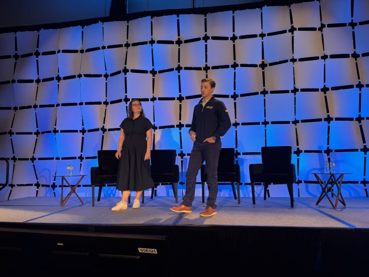 Two people standing on a stage. One person is talking to an audience with their hands in their pockets and the other person is looking at them.