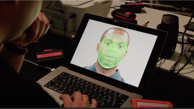 A person with their back to us sits at a computer. On the screen is an image of a black man's face. Superimposed over his face is a green neon grid pattern mapping the contours and movements of his face.