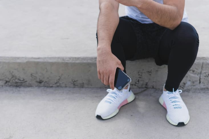 A close-up shot from the chest down of a person sitting on a concrete ledge, wearing black pants and white athletic shoes with pink accents. Their right hand rests on their leg while holding a dark-colored smartphone.