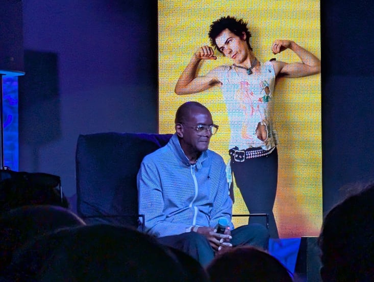 Dennis Morris, a black man in glasses and a blue zip up jersey sits in front of a screen showing his photograph of Sid Vicious from the Sex Pistols.