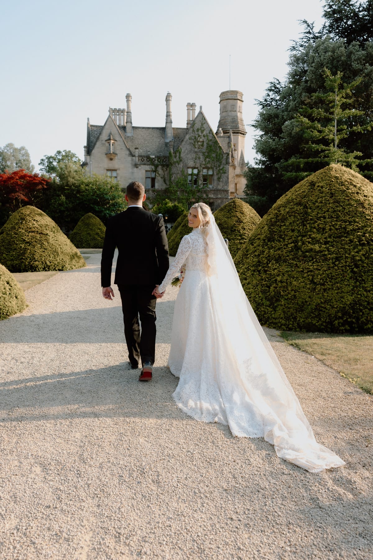 Bride and groom walking hand in hand towards a Cotswolds manor house, her veil trailing behind