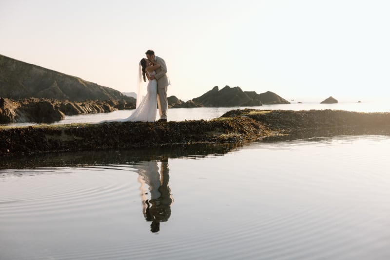 Bride and groom on the rocks at Tunnels Beaches, Devon, reflected in the still water