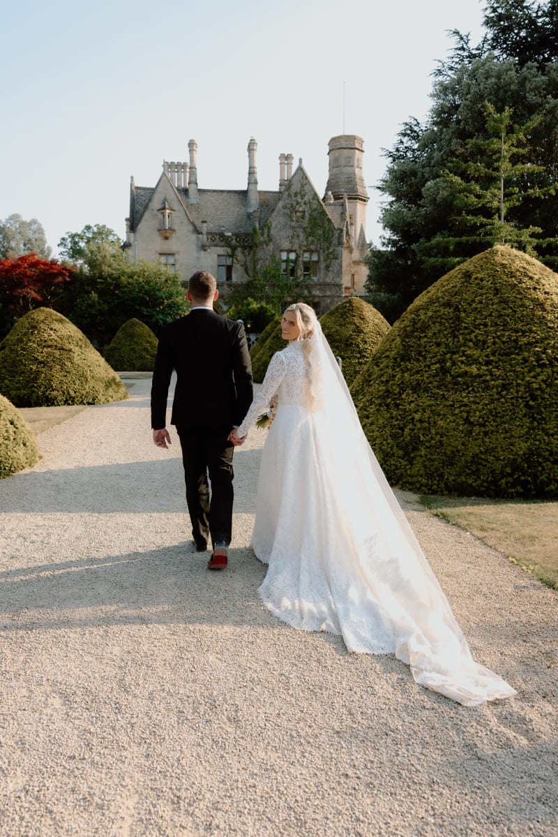 Bride and groom walking towards the manor house, her train trailing behind, The Cotswolds