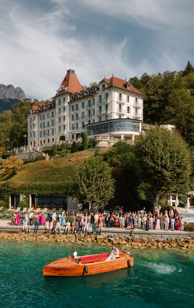 A wooden boat on Lake Annecy below the Palace de Menthon, guests gathered along the quayside