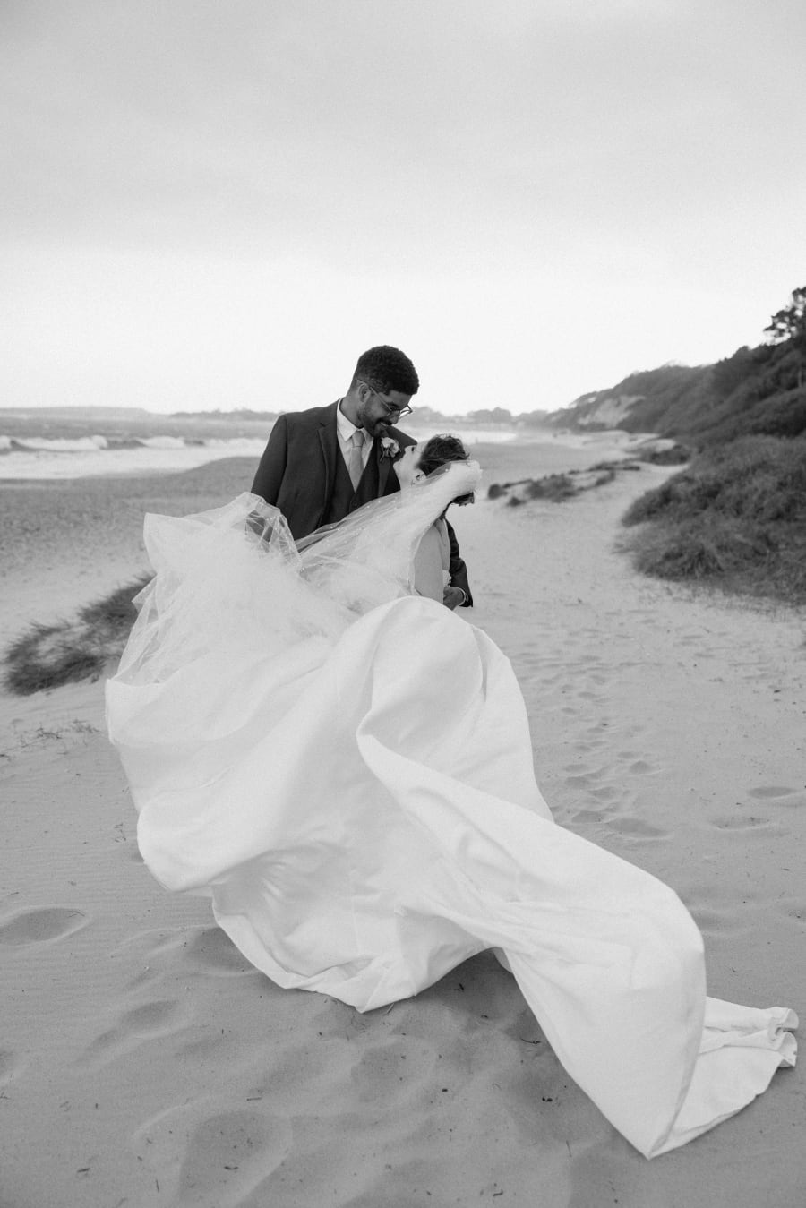 Her dress caught by the wind as they hold each other on the sand, in black and white, Highcliffe Castle, Christchurch