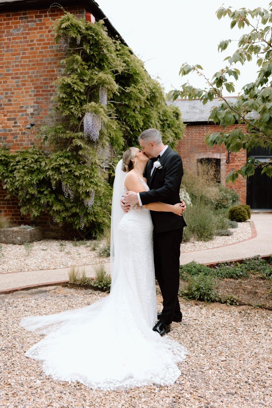 Amy and Matt holding each other beside the ivy-covered brick barn, Clock Barn, Whitchurch