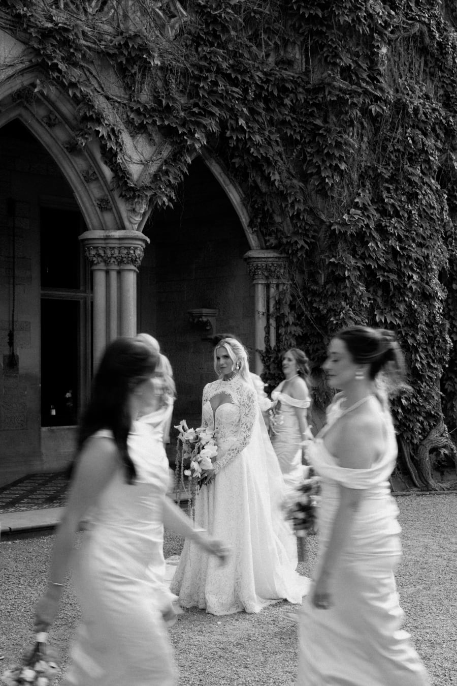 Bridesmaids passing the bride outside the ivy-covered archway, in black and white, The Cotswolds