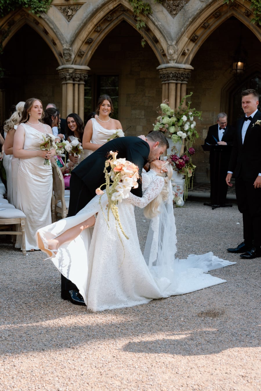 Josh dipping Morgan for a kiss in the cloisters as the bridal party look on, The Cotswolds