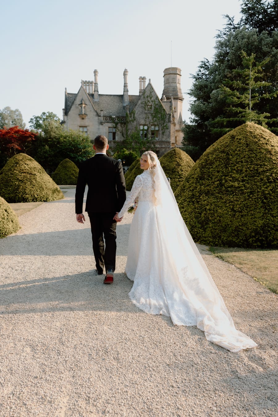 Morgan and Josh walking towards the manor house between the topiary, her train trailing behind, The Cotswolds