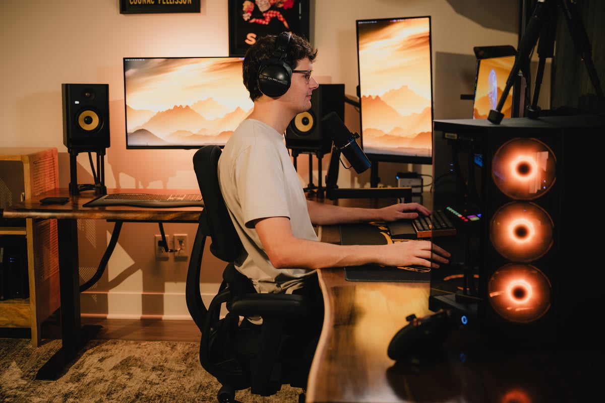 Side-profile view of a person seated in the Anthros chair at a multi-monitor workstation, elbows resting near 90 degrees with forearms parallel to the desk surface, demonstrating correct desk height and posture support.