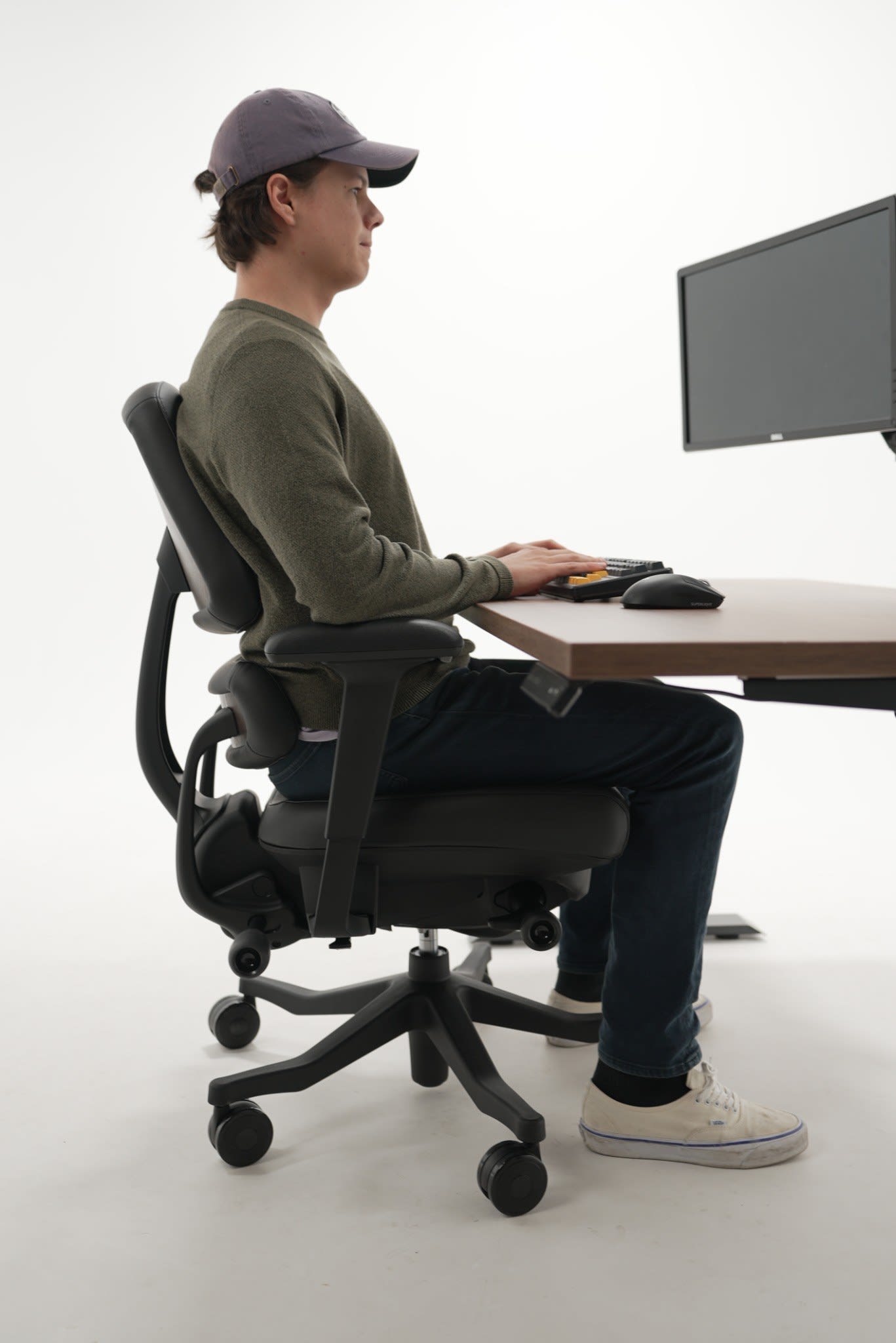 Left side-profile of a male model seated in the Anthros chair at a standing desk, typing against a white studio backdrop, demonstrating a relaxed, aligned posture supported by the chair's backward tilt.