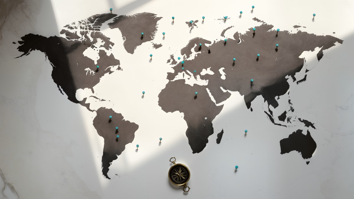 Black ink world map on white marble with teal pins in 76 countries and brass compass, showing global reach of 5,000+ Cracking Superhost students