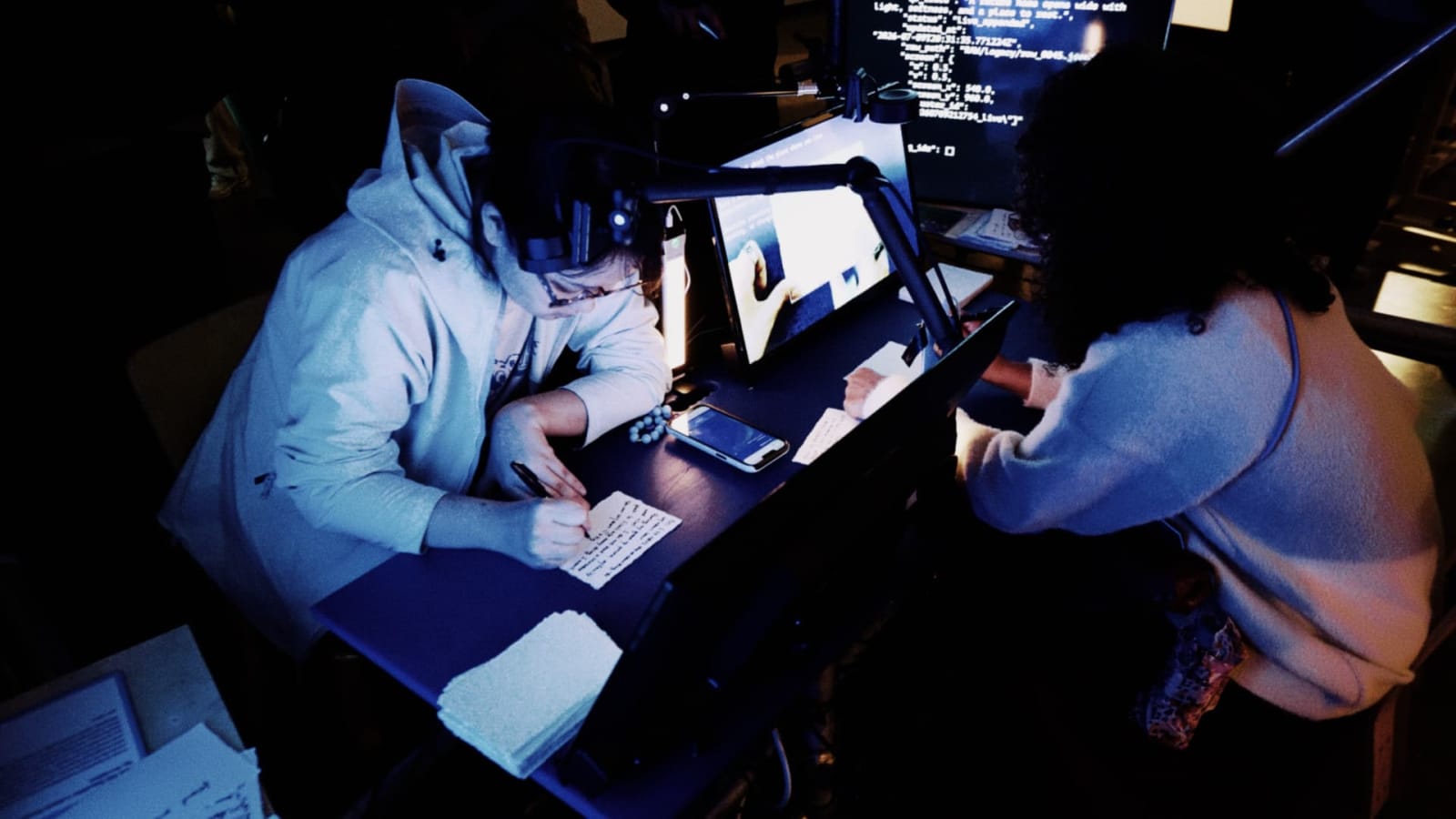 Two people sit together at the writing station, with paper on the desk and a screen in front of them.