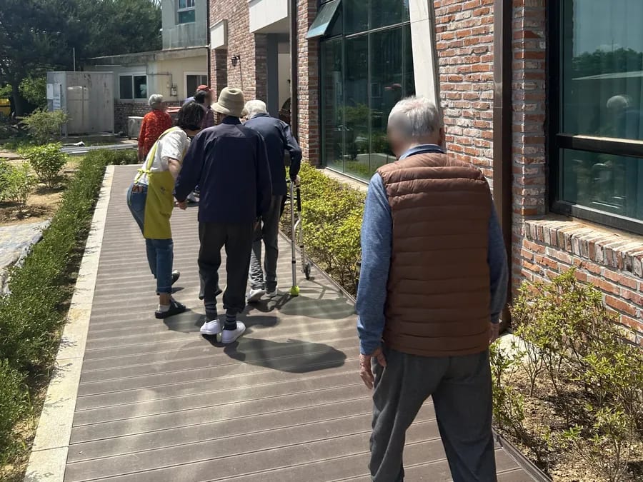 벽돌 건물 옆 데크 길을 걸어가는 어르신과 직원들의 뒷모습