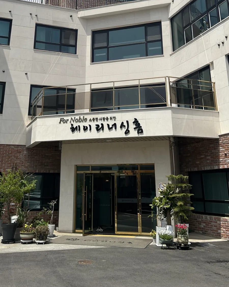 벽돌과 흰 벽의 3층 건물 정문 — 발코니의 'For Noble 소중한 사람을 위한 헤이리너싱홈' 간판, 금색 테 유리문, 정문 앞 화분