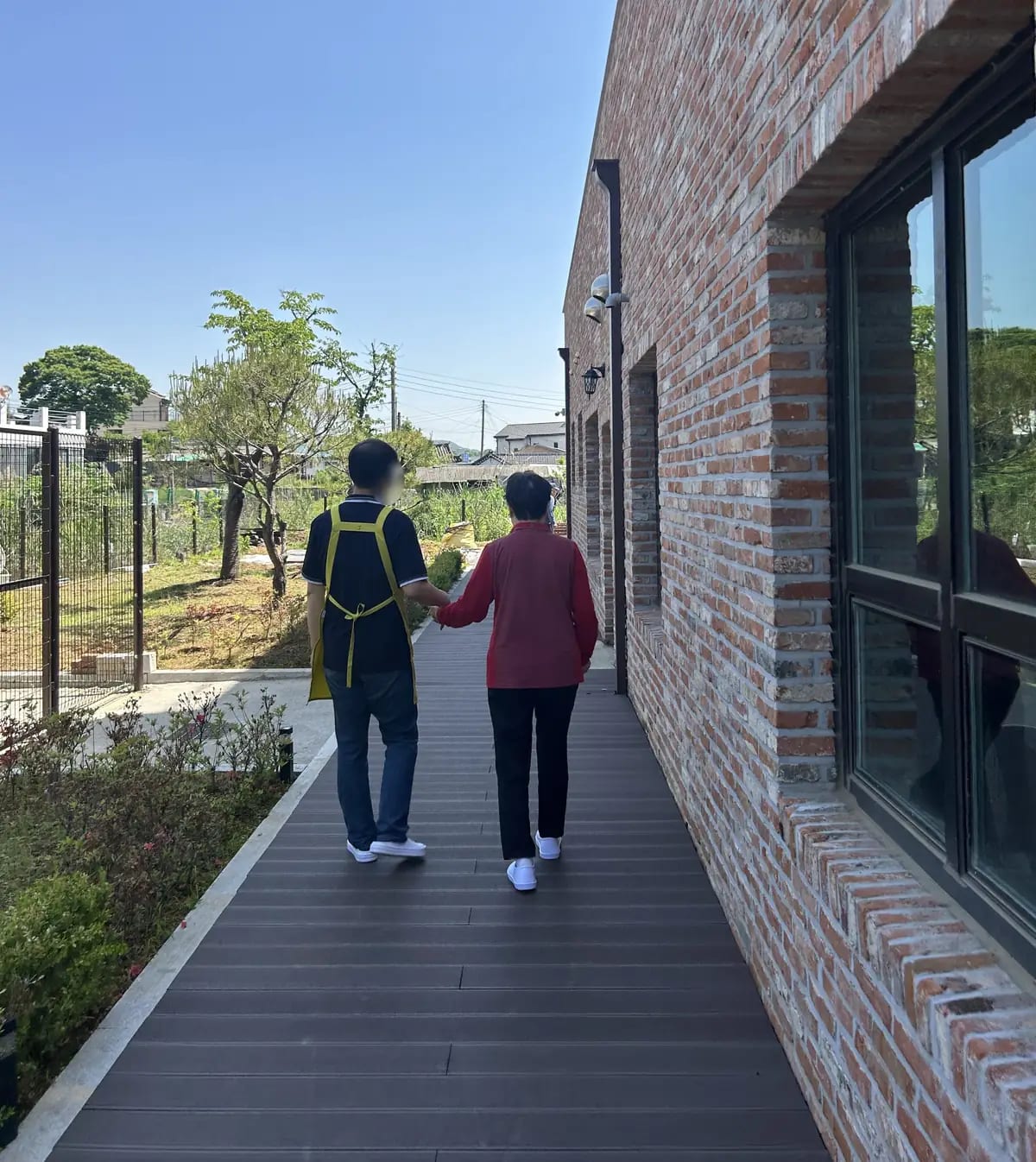 벽돌 건물 옆 나무 데크에서 앞치마를 두른 직원이 어르신 손을 잡고 함께 걷는 뒷모습 — 맑은 하늘과 텃밭