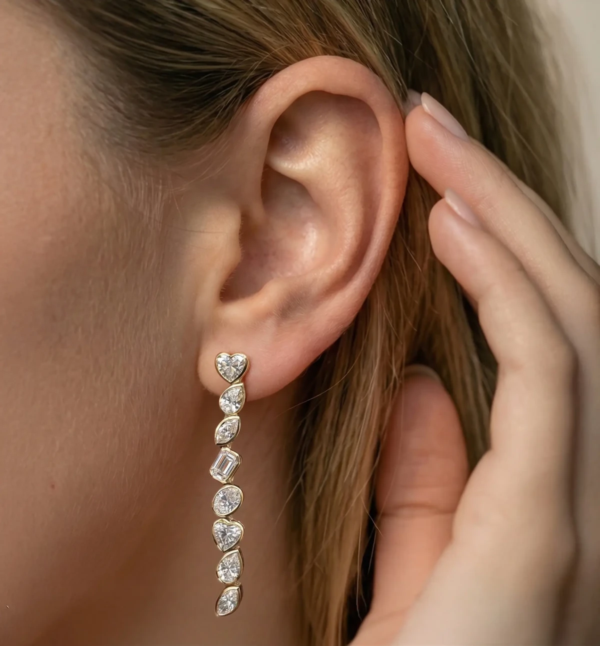 A long drop earring of pear and emerald-cut diamonds on a woman's ear, her fingers at her hair