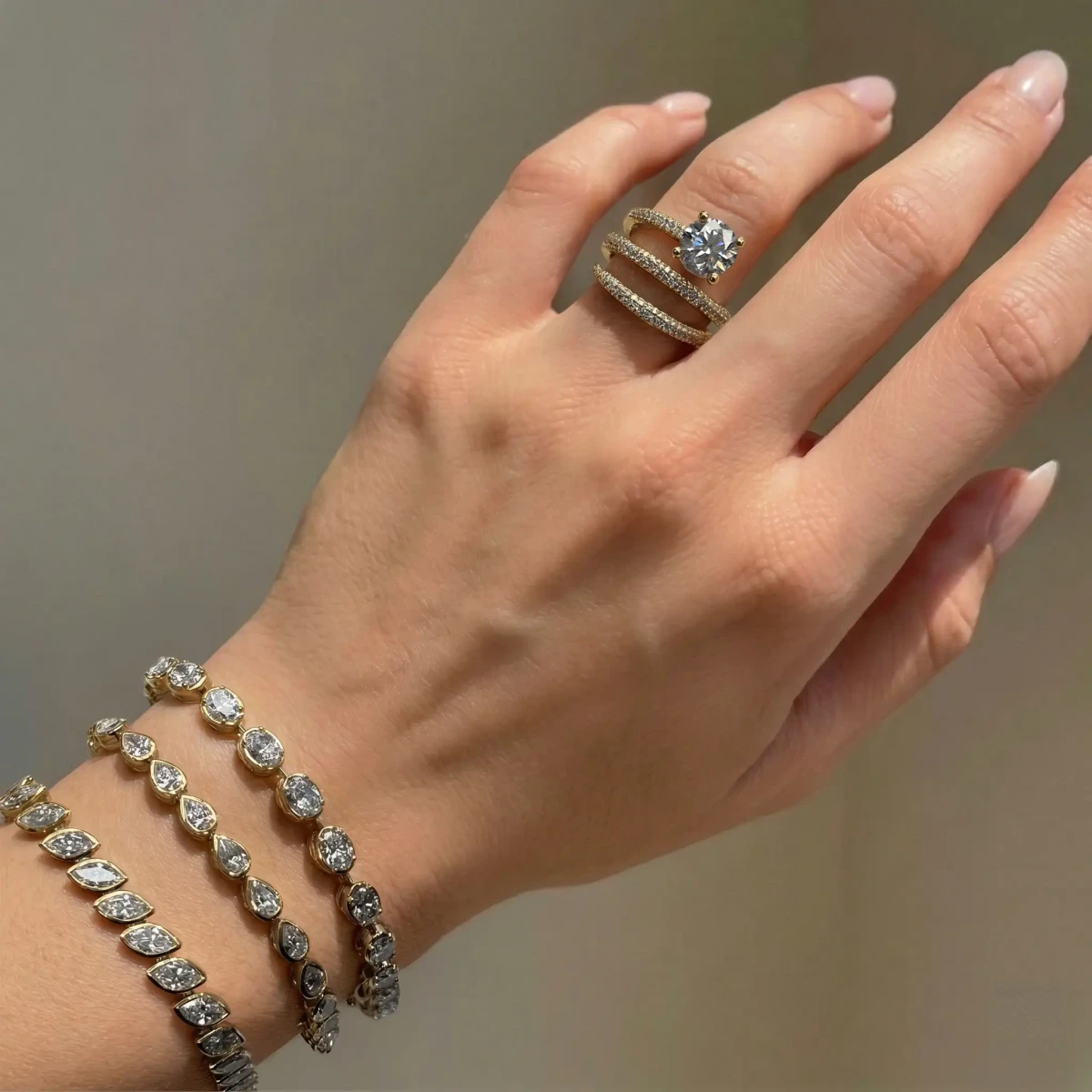 A hand wearing a gold ring with a round diamond and three gold diamond bracelets, against a warm wall