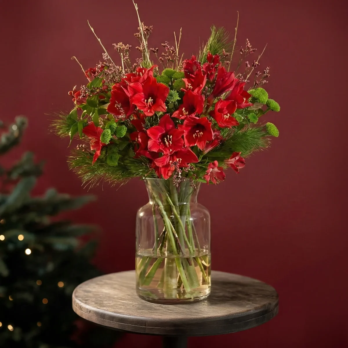 A bouquet of red amaryllis and greenery in a glass vase on a small wooden table, a Christmas tree soft in the background