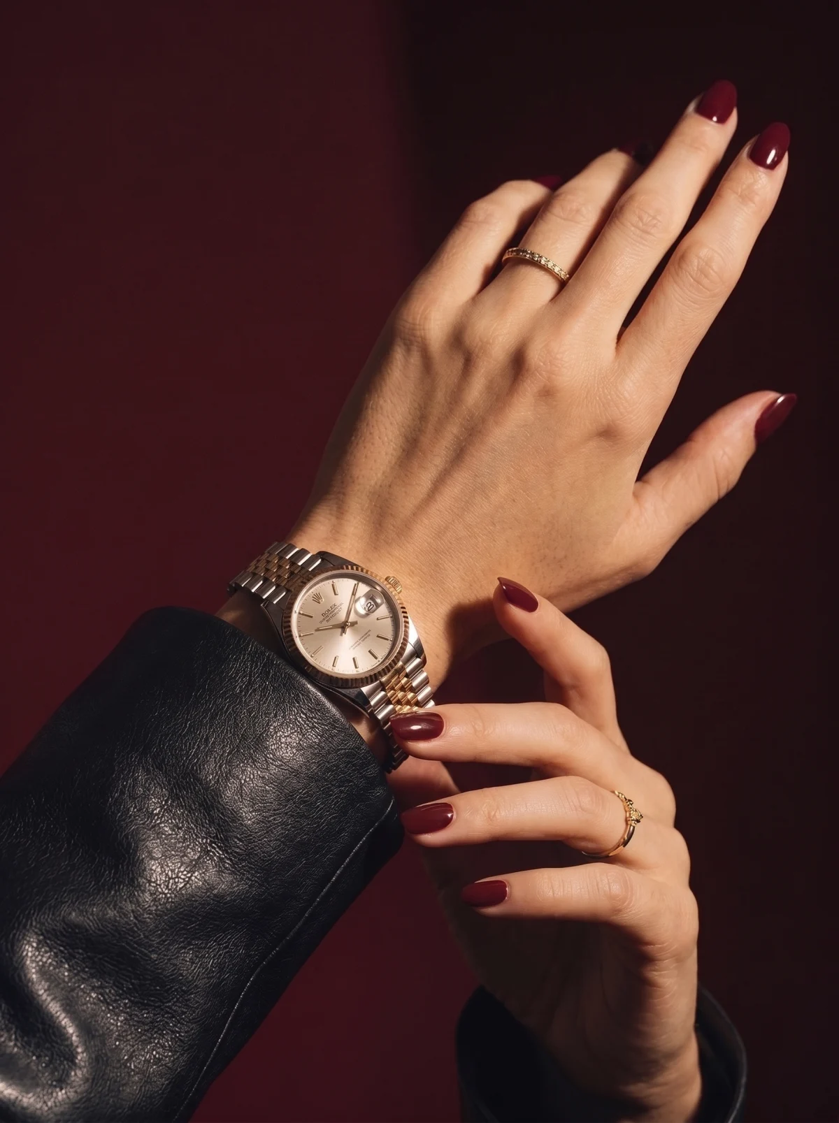 A two-tone steel and gold watch on a woman's wrist, dark leather sleeve, burgundy background