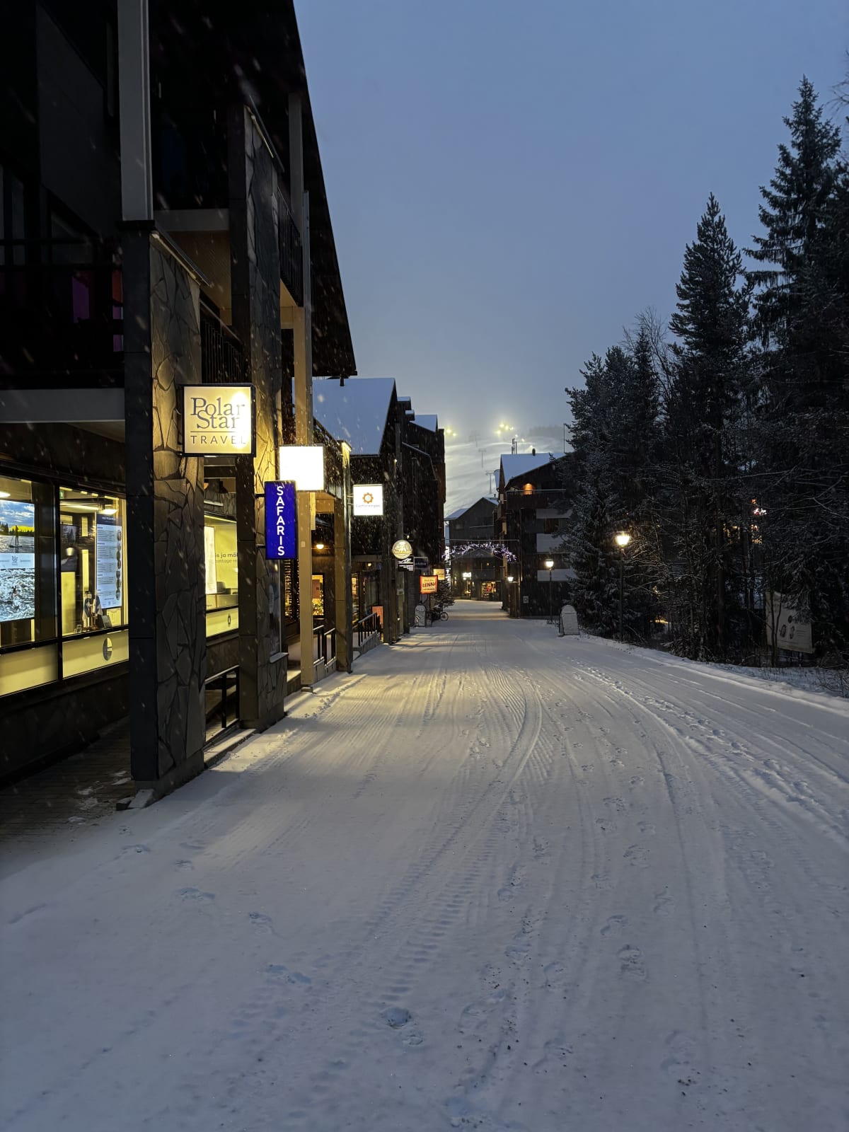 A snowy street through Levi village at dusk, shopfronts lit along it