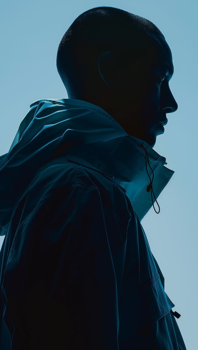 Silhouette of a person in a dark hooded jacket in profile against a pale blue background