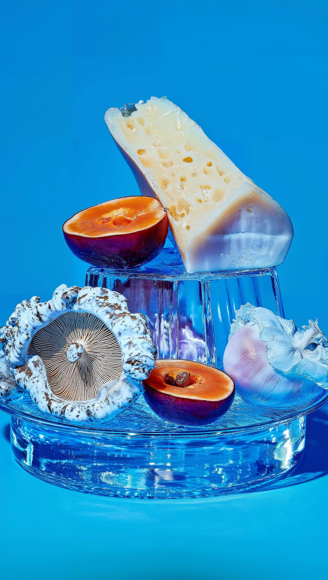 Still life of cheese and halved plums on a blue glass stand against a saturated blue backdrop