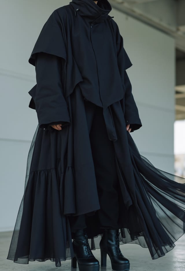 Person in a long dark coat and tiered tulle skirt with platform boots, skirt moving in the wind