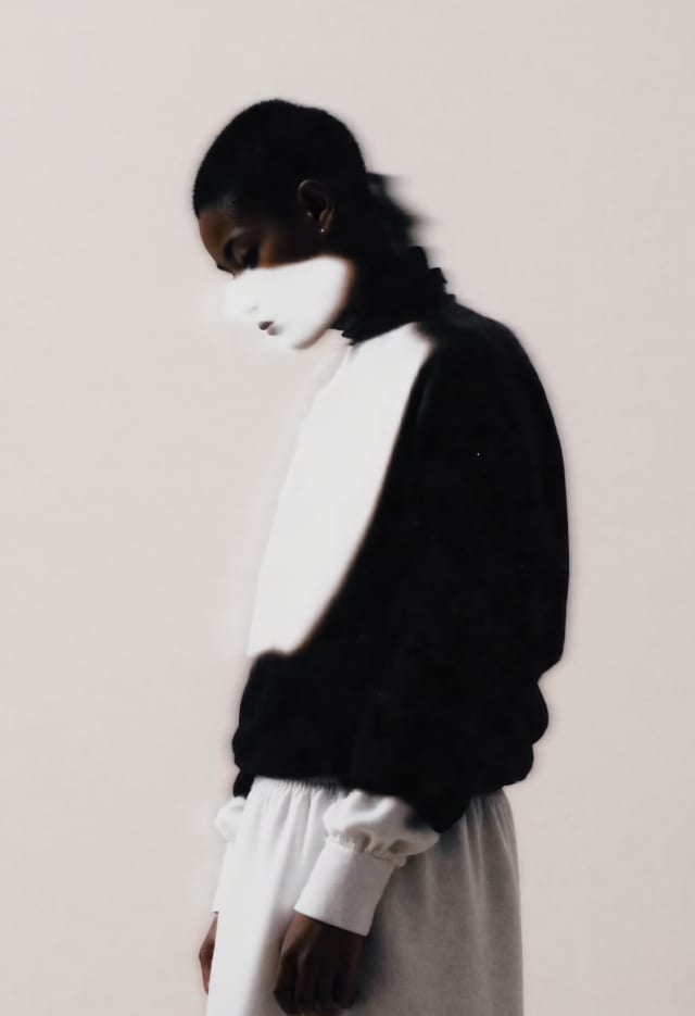 Blurred profile of a person in a black and white knit with a white face mask against a pale wall