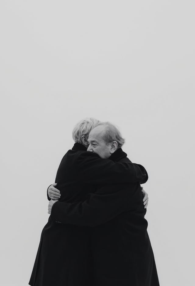 Two people in black coats hugging against a light grey background