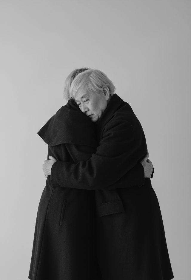 Two people in dark coats in a tight embrace, one with silver hair, on a pale background