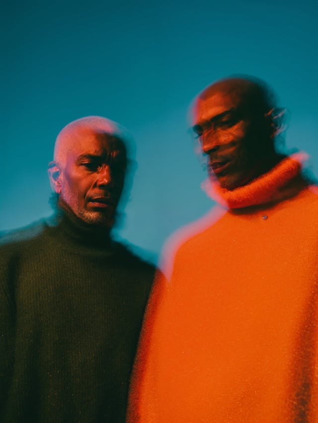 Two men in a turquoise light, one in a dark green turtleneck, one in a glowing orange sweater, blurred motion