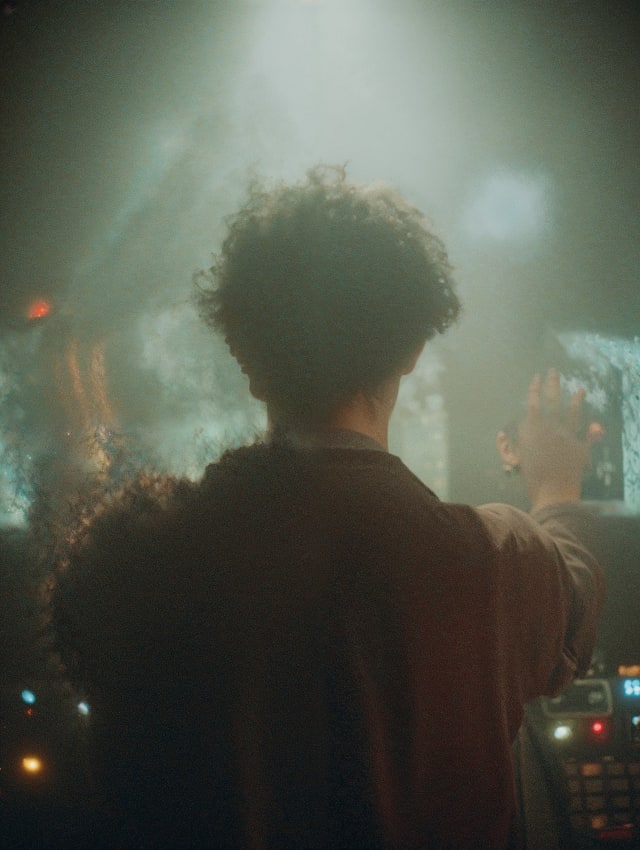Curly-haired person seen from behind in a hazy, light-filled room with a screen glowing in the background