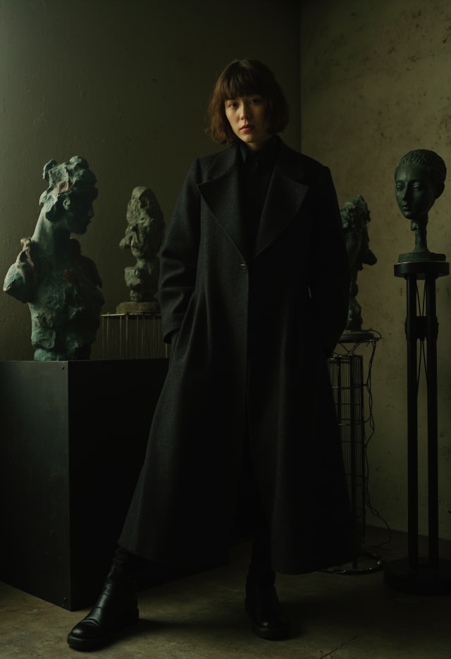 Woman in a long black coat standing among sculptures in a dim room