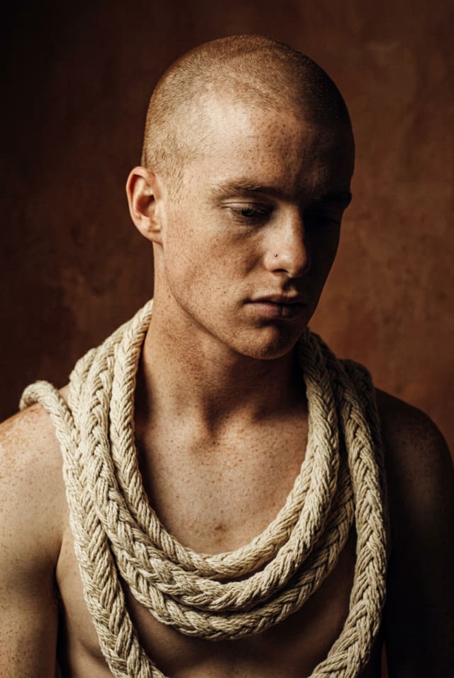 Shaven-headed man with a thick rope coiled around his neck against a warm brown background