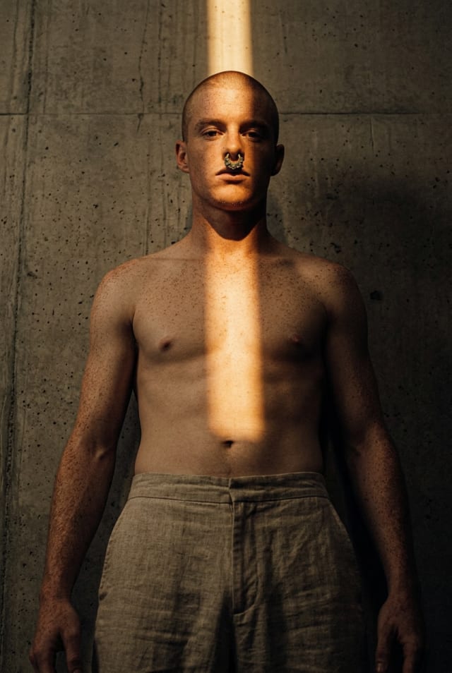 Shirtless man with a nose ring standing in a concrete room, a shaft of light across his torso