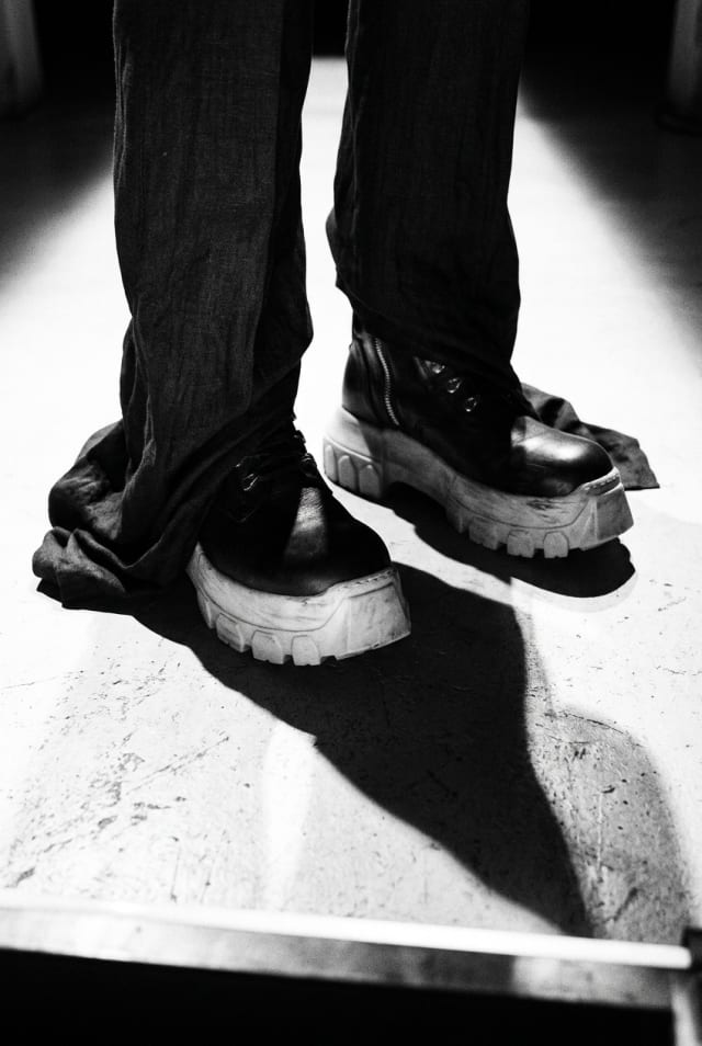 Black and white shot of chunky black boots and dark trousers casting a hard shadow