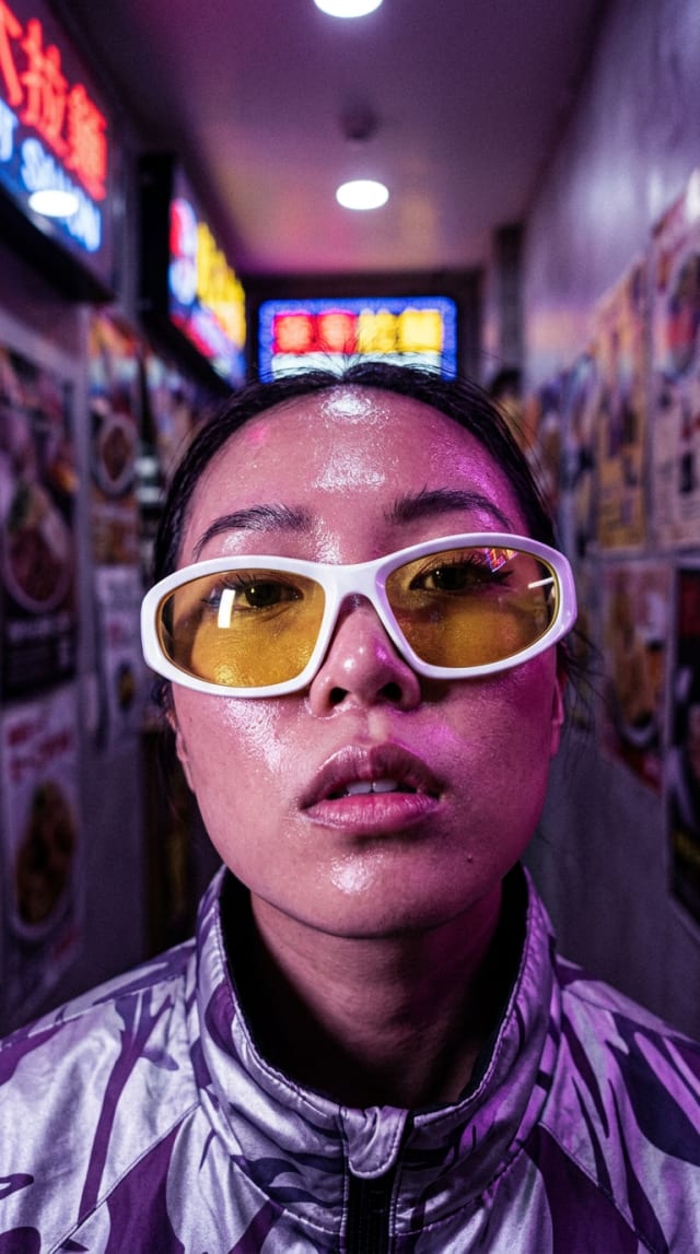 Portrait of a woman in a metallic violet jacket and yellow-tinted white sunglasses in a neon-lit alley