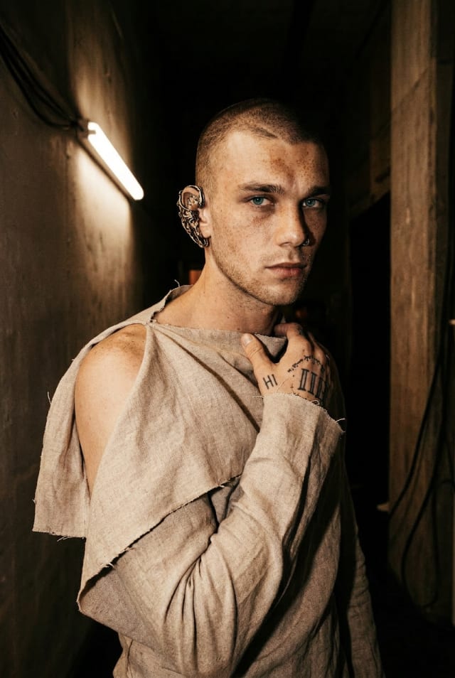 Man in a torn beige linen top standing in a narrow dark corridor under a strip light