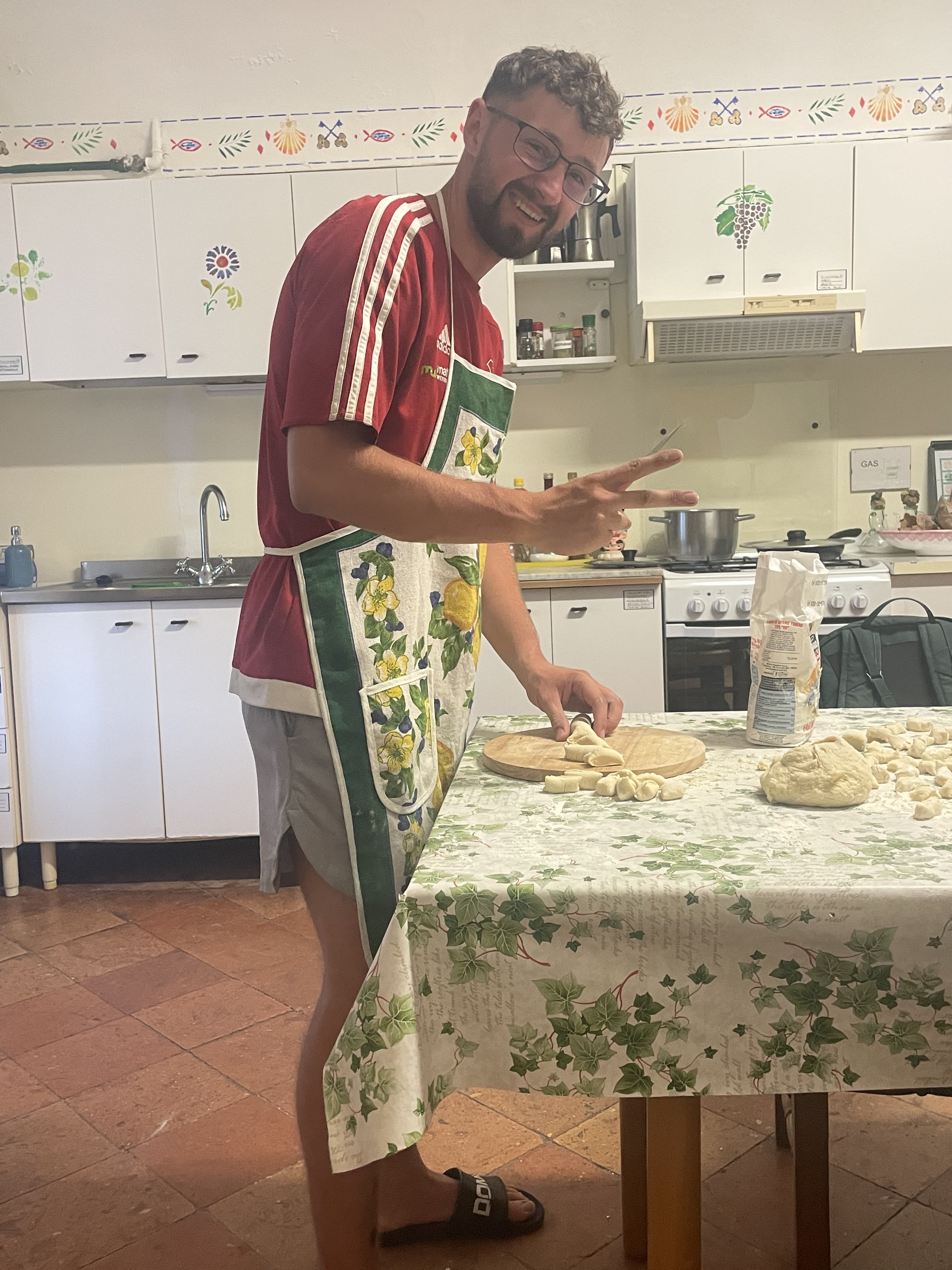Steven in a floral apron making gnocchi from scratch in the pilgrim house kitchen – flour on the table, peace sign, huge smile