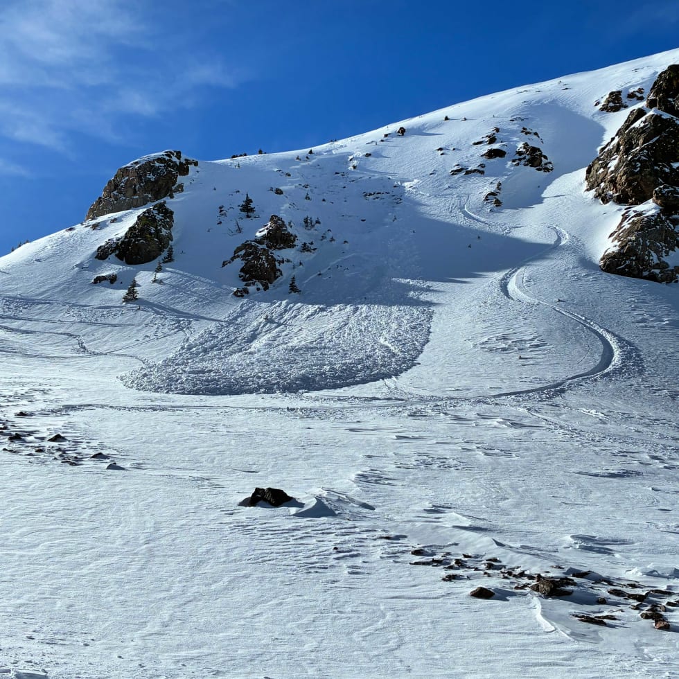 Skier-triggered slab avalanche on a steep alpine slope in the Northern Mountains, showing a defined crown, debris pile at the base, and ski tracks adjacent to the slide path under a clear blue sky.