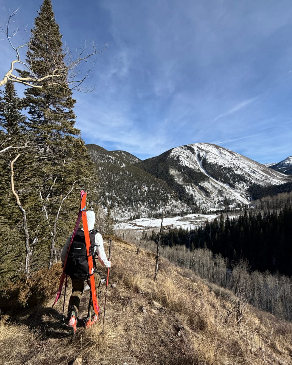 Backcountry forecaster carrying skis hikes down a dry, south-facing ridge near Ashcroft in the Elk Mountains, with patchy snow on surrounding slopes and bare ground at about 10,500 feet.