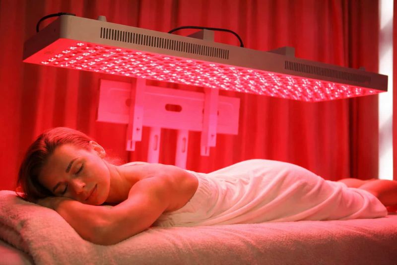 Woman undergoing red light therapy treatment under a red LED panel in a relaxing wellness atmosphere