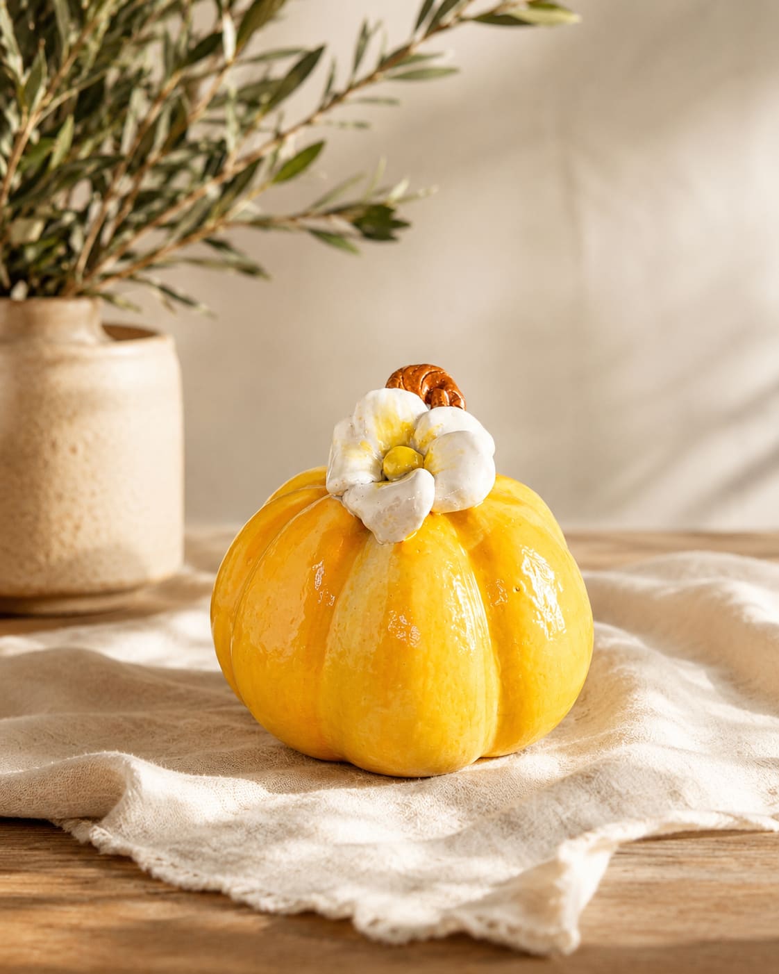 Handmade ceramic pumpkin with golden-yellow glaze and a white sculpted flower on top, placed on a linen cloth