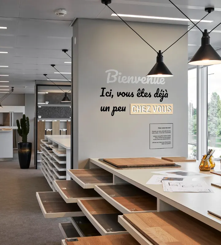 Photo du showroom Kaufman Broad de Courbevoie avec différentes palettes de matériaux pour personnaliser votre futur logement et la phrase inscrite sur un mur :
"Bienvenue 
Ici, Vous êtes déjà un peu chez vous"