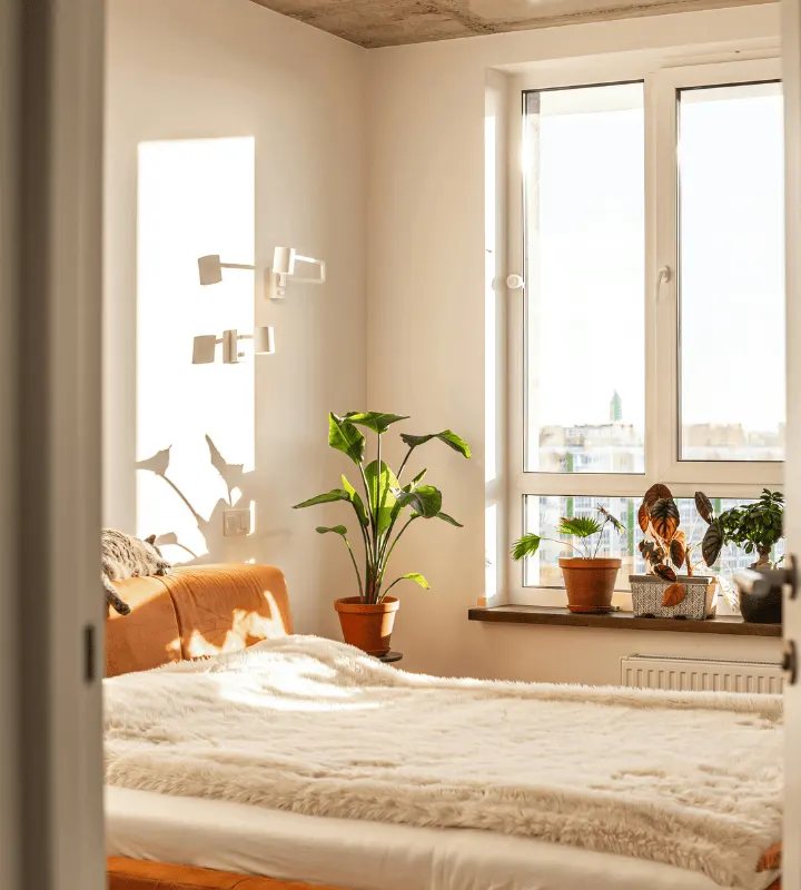 Chambre moderne baignée de lumière naturelle, décorée dans des tons clairs et chaleureux. Un grand lit recouvert d’une couverture beige est placé près d’une grande fenêtre laissant entrer le soleil. Quelques plantes vertes et pots en terre cuite apportent une touche naturelle et apaisante. L’ambiance générale est douce, cosy et minimaliste