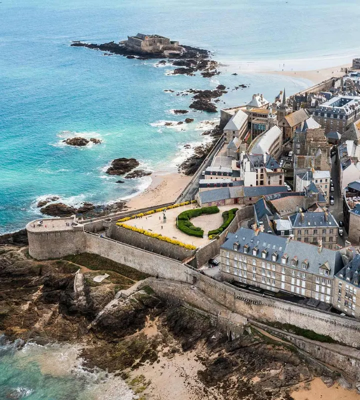 Vue aérienne d’un quartier historique en bord de mer, mettant en valeur une architecture traditionnelle avec des toits en ardoise, entourée de remparts en pierre. L’ensemble est situé face à l’océan, avec une plage de sable fin et des rochers. La présence d’un fort sur un îlot en arrière-plan renforce le caractère patrimonial du lieu. Cette image reflète un cadre de vie authentique et prestigieux, entre patrimoine architectural et panorama maritime.