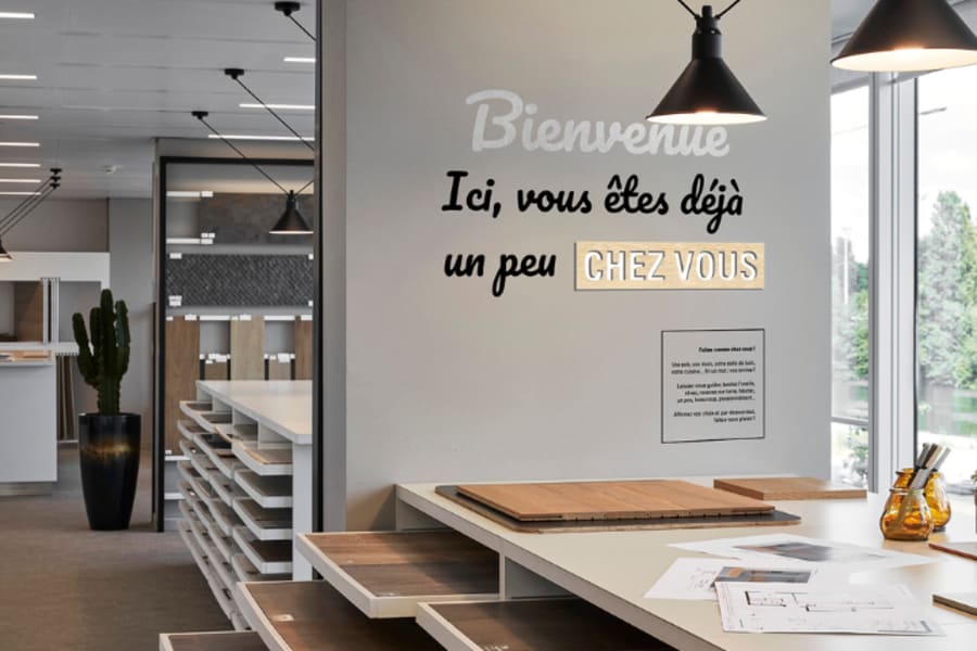 Photo du showroom Kaufman Broad de Courbevoie avec différentes palettes de matériaux pour personnaliser votre futur logement et la phrase inscrite sur un mur :
"Bienvenue 
Ici, Vous êtes déjà un peu chez vous"
